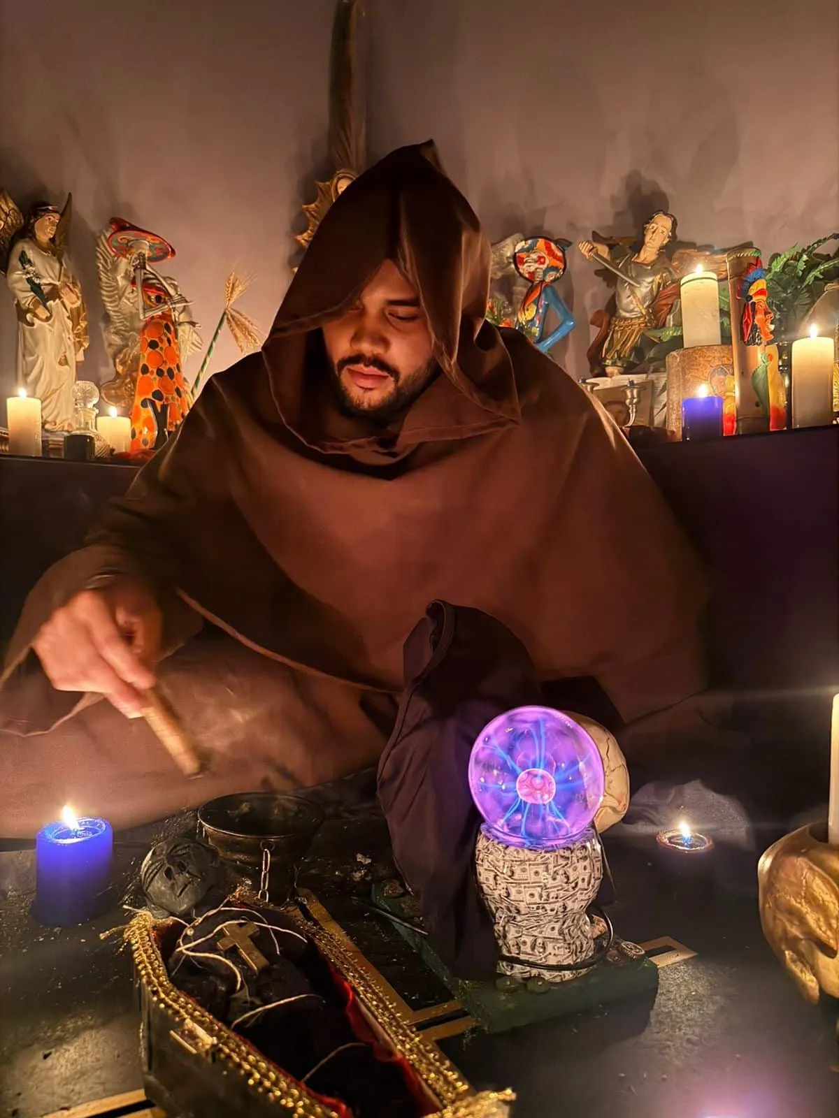 El Maestro Abel con hábito marrón y capucha, preparando un ritual frente a su altar de santos y velas